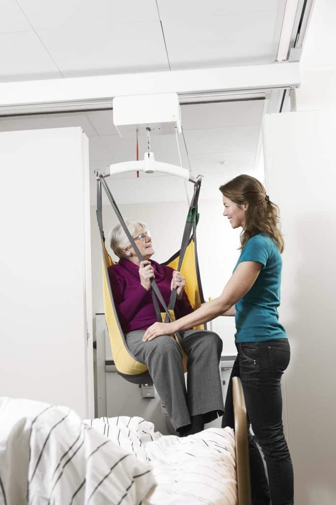 An elderly woman in a yellow sling Guldmann GH3 Ceiling Lift assisted by a woman in a teal shirt.