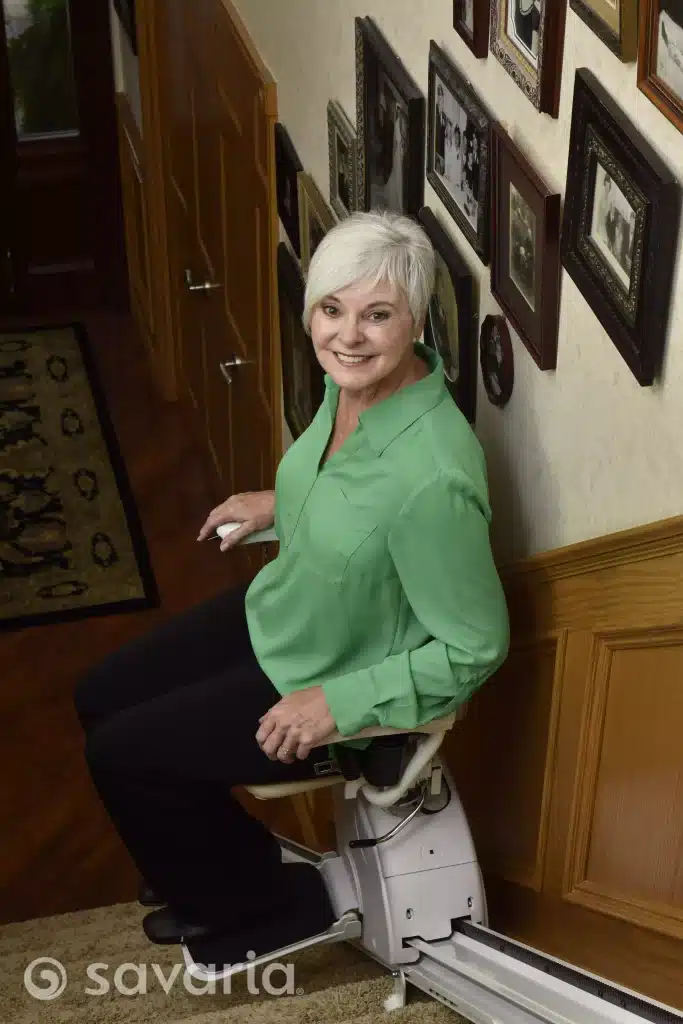 An older woman in a green shirt sits on a Savaria-K2-stairlift-model, smiling, with framed photos on the wall behind her.