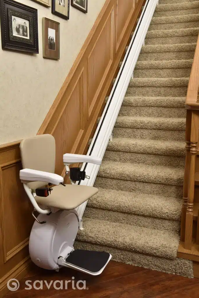 Savaria K2 stairlift unfolded installed on a carpeted staircase in home with wooden paneling and framed photos above on the wall.