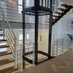 Savaria Vuelift Glass Elevator in Modern Home, Interior surrounded by a wooden staircase with metal railings.