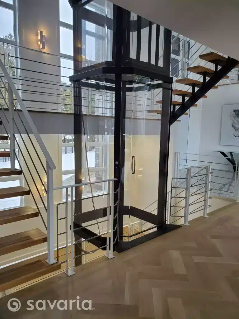 Savaria Vuelift Glass Elevator in Modern Home, Interior surrounded by a wooden staircase with metal railings.