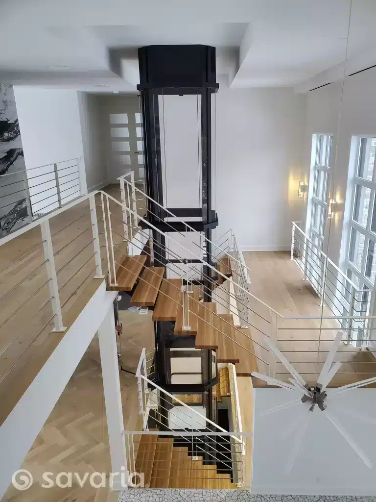 Savaria Vuelift Glass Elevator, Interior view of a modern space with a central glass elevator and spiraling wooden staircase.