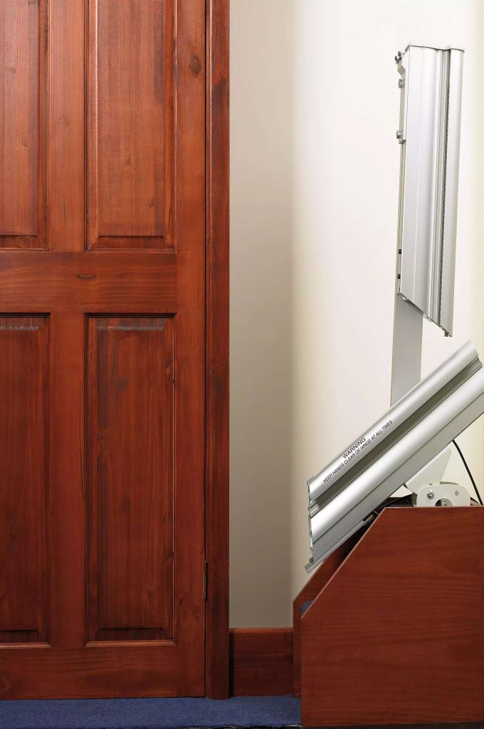 Wooden door next to a metallic Handicare 1000 straight stairlift on a cream-colored wall.