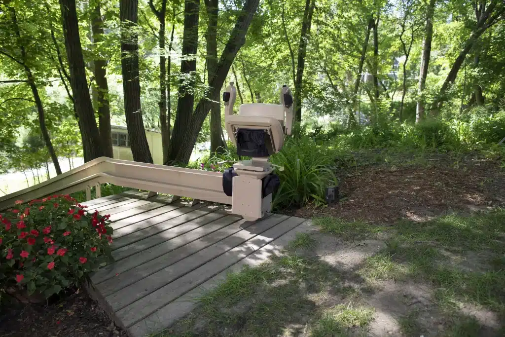 Bruno Elite Outdoor Curved Stair lift on a wooden deck in a lush green outdoor setting with red flowers.