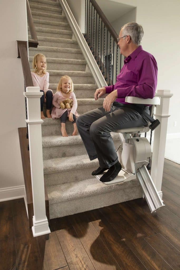 An elderly man sitting on a Bruno Elite Straight Rail Stairlift, interacting with two young girls on a carpeted staircase, showcasing Access and Mobility home accessibility solutions for seniors.