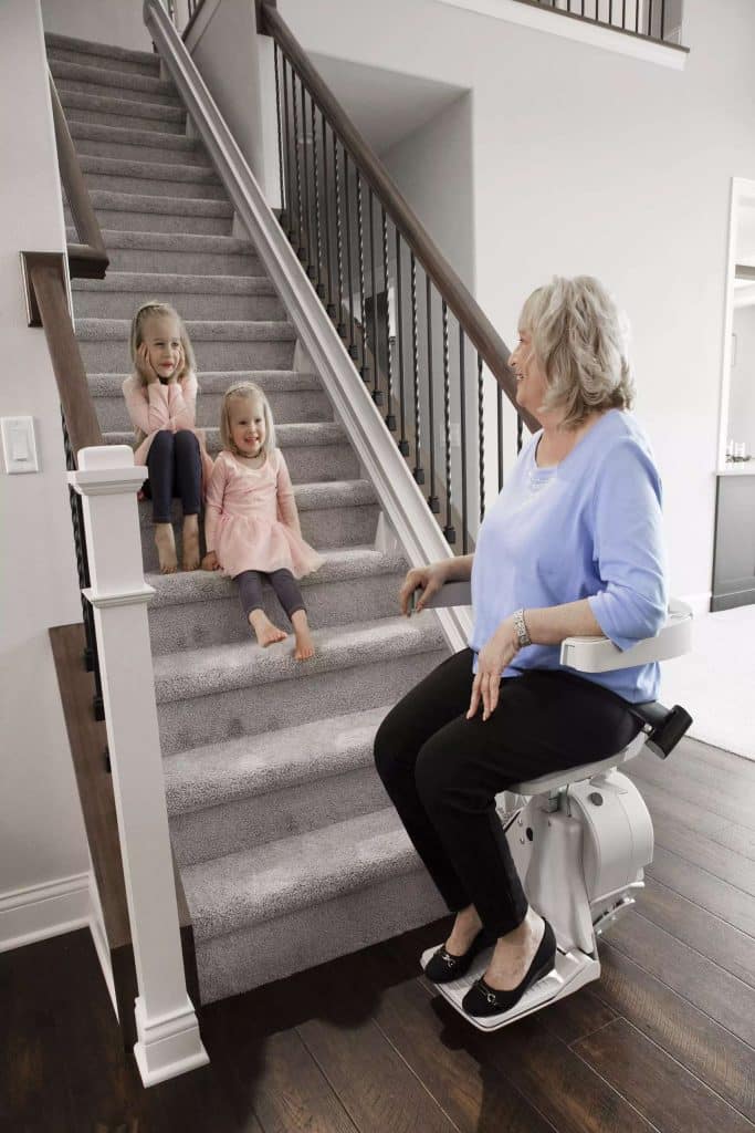 An elderly woman sitting on a Bruno Elite Straight Rail Stairlift, interacting with two young girls on a carpeted staircase, showcasing Access and Mobility home accessibility solutions for seniors.