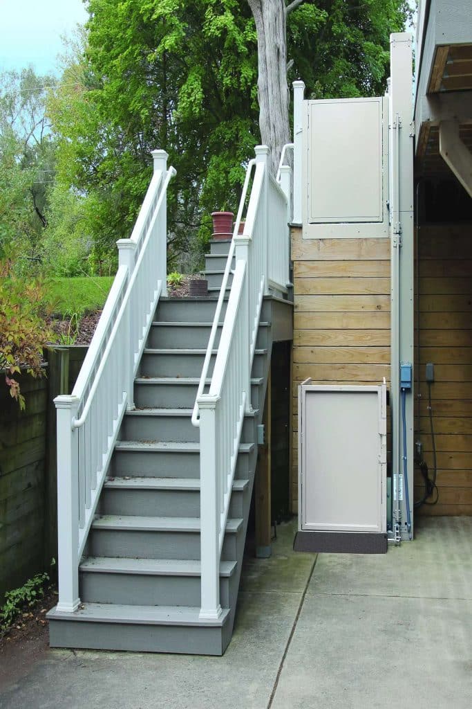 Outdoor Bruno Vertical Platform Lift next to gray stairs with white railings, offering mobility access to an elevated deck area.