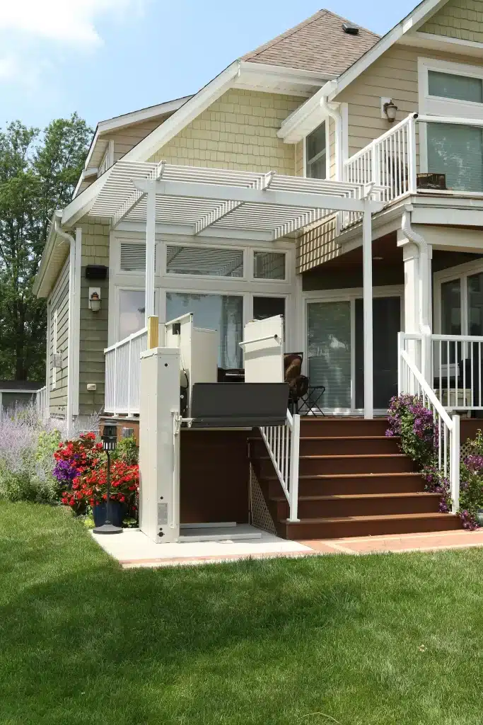 Exterior of a suburban house with a white Bruno Vertical Platform lift installed next to wooden stairs leading to a deck with a pergola. Surrounded by a green lawn and vibrant flowers.