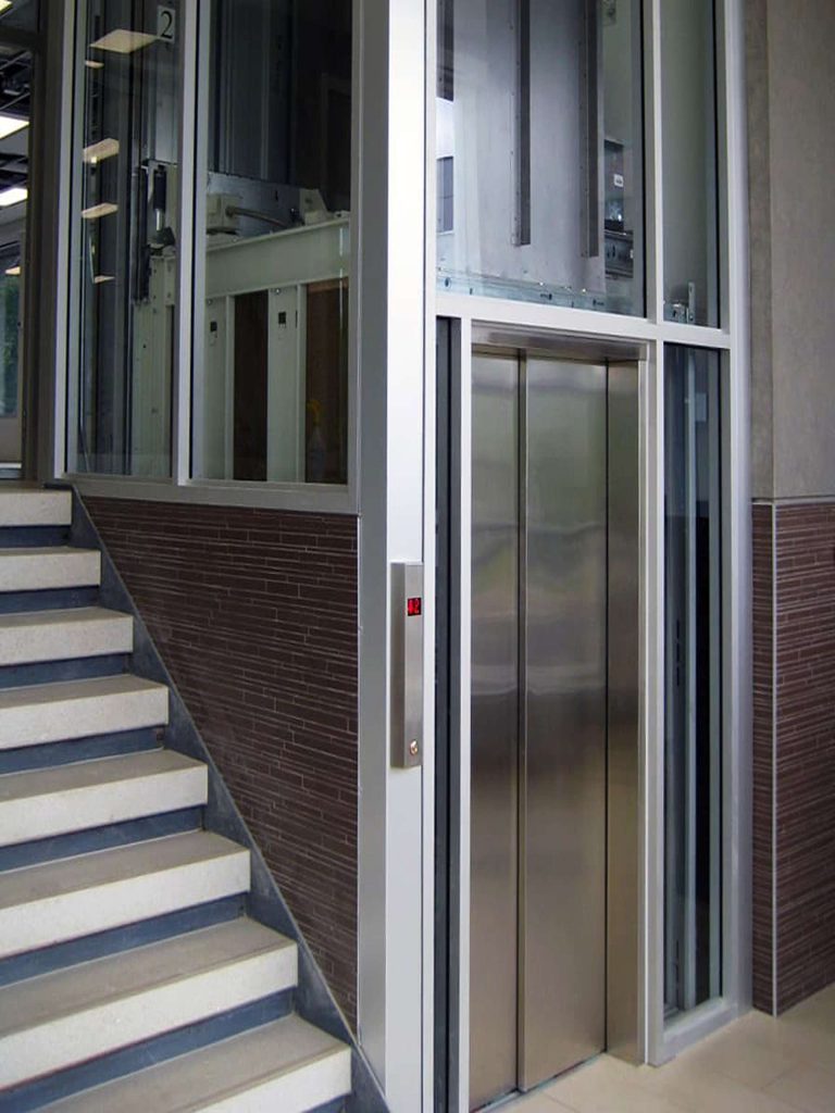 Savaria Orion Commercial Modern Glass Elevator with Stainless Steel Doors Next To Staircase in Building Interior.