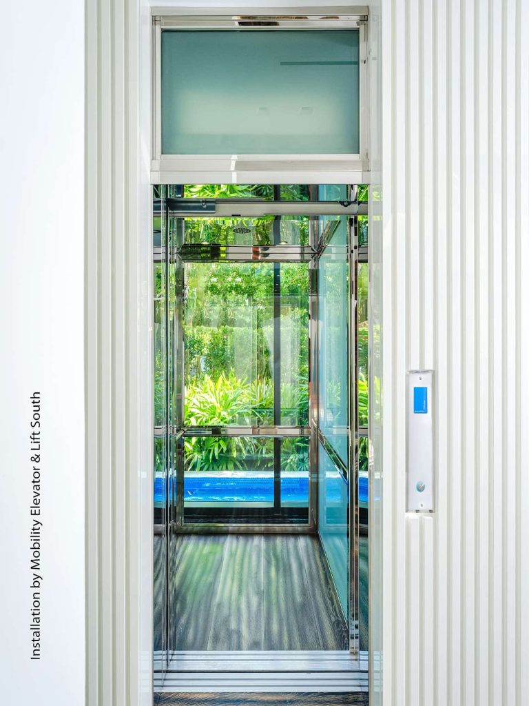 Savaria Orion Commercial Transparent Elevator With Glass Panels and Metal Interior set against White Ridged Walls with Greenery Visible Outside.