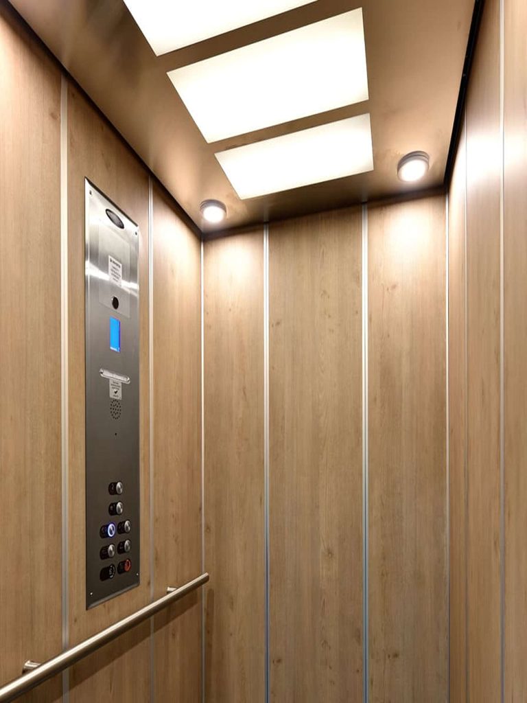 Interior of Savaria Orion Commercial Wood Paneled Elevator with Metal control panel and lighted ceiling.