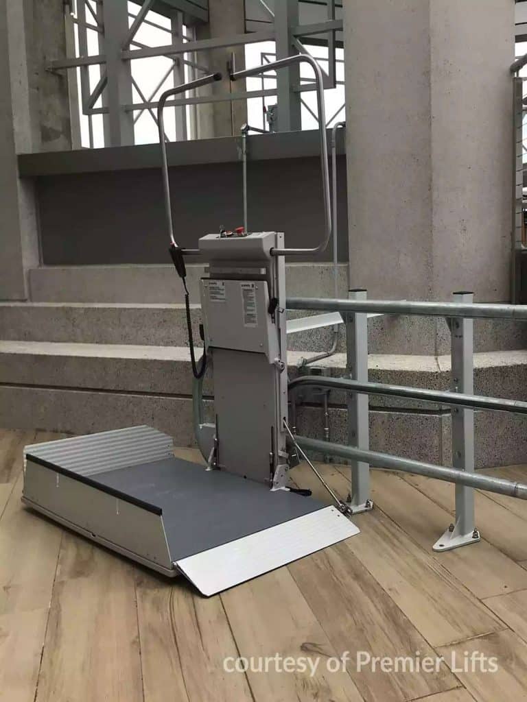 Savaria Omega Inclined Platform lift system installed next to concrete stairs with metal railings and wood-textured flooring.