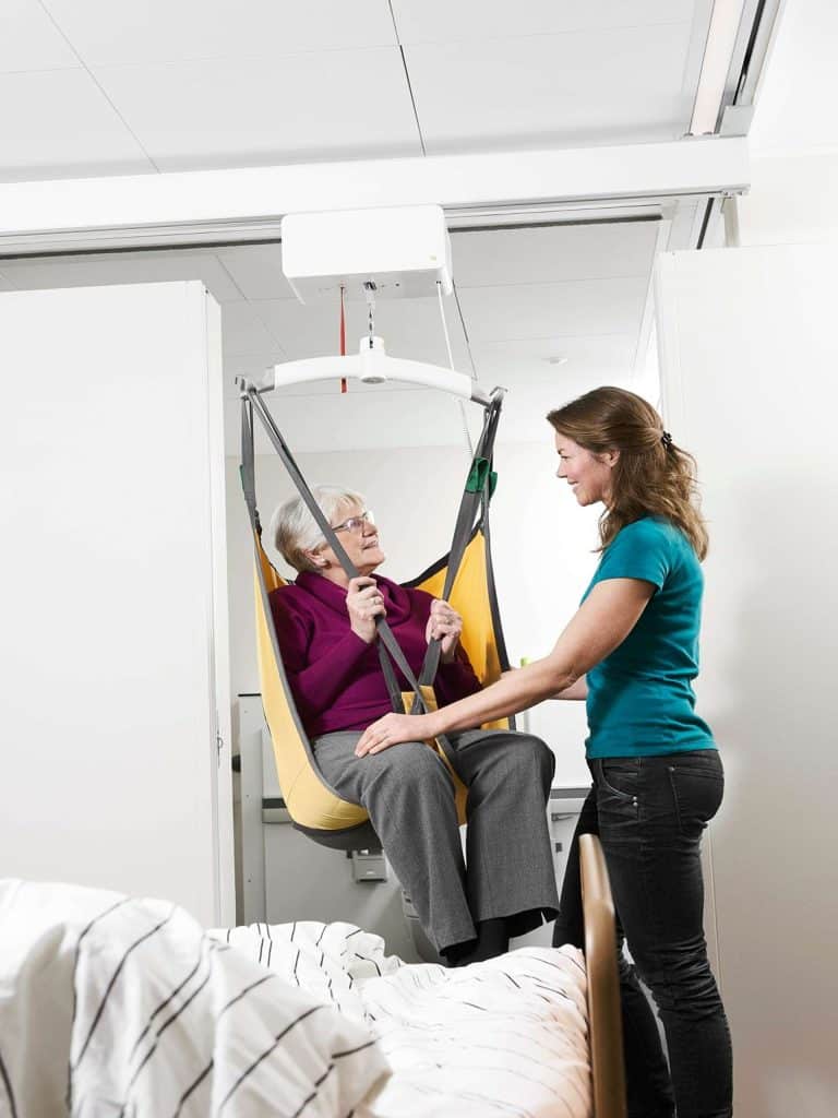 An older adult seated in a yellow sling Guldmann GH3 Ceiling Lift, supported by a caregiver next to a bed.