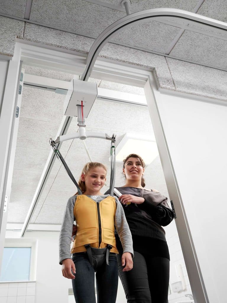 A girl in a yellow harness is supported by a Guldmann GH3 Ceiling mounted track system with woman assisting from behind.