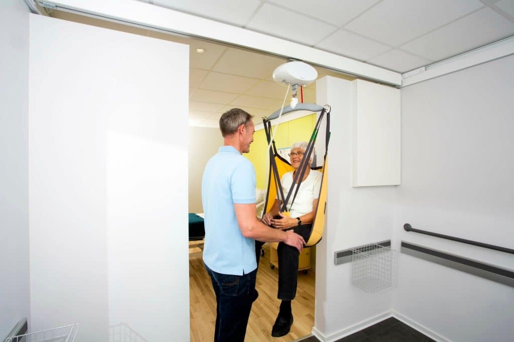 A man assists an elderly woman with Guldmann GH1 Ceiling Lift System in a care facility.