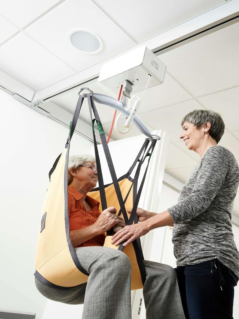 A woman assists an elderly woman seated in a yellow lift sling attached to a Guldmann GH3 ceiling track lift system.