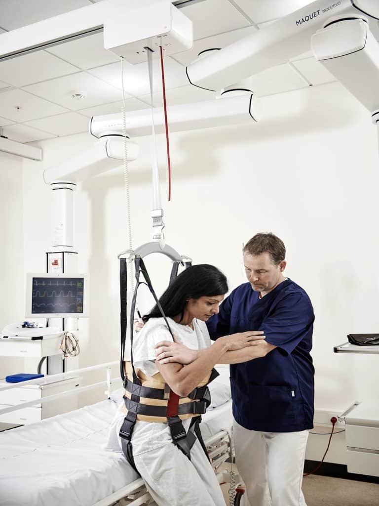 Healthcare professional assists a woman in a harness connected to a Guldmann GH3 ceiling track lift system in a medical room.