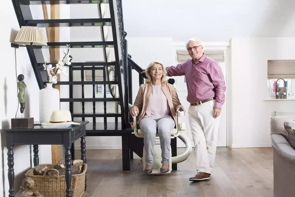 An elderly couple in a cozy living room with the woman sitting on a Handicare Freecurve Curved stairlift emphasizing comfort and accessibility in a home environment.