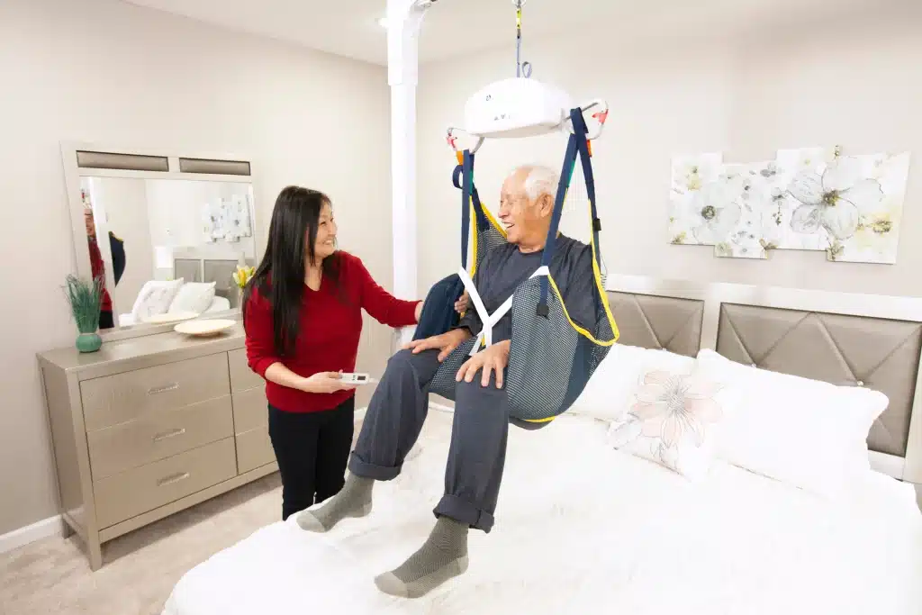 An elderly person is in Handicare AP Series Portable Ceiling Lift over a bed assisted by a smiling woman with a remote in a bright bedroom.