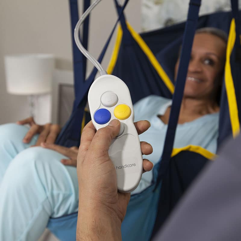 Close-up of a hand holding a remote control of Handicare C-450 ceiling lift with four buttons against a background of a person in a hoist sling.