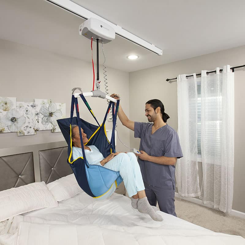 A healthcare worker using an Handicare C-450 ceiling lift system to assist person in a blue harness above a bed in a bedroom.