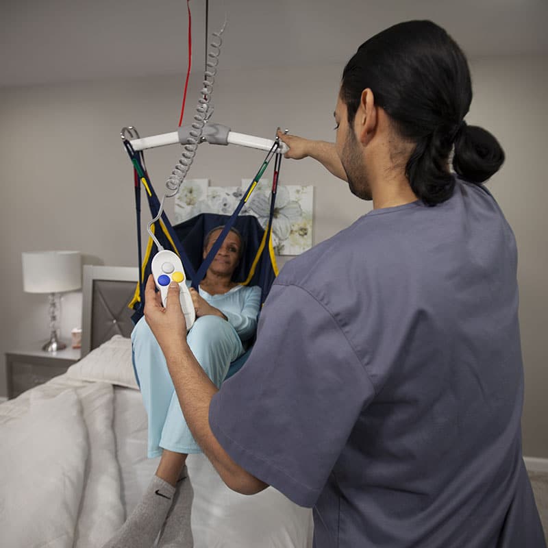 Caregiver using Handicare C-450 ceiling lift to assist a woman seated in a sling in a bedroom.