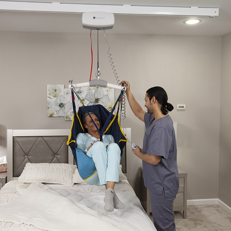 Caregiver operating a Handicare C-450 ceiling mounted lift system, lifting a woman from a bed in a blue and yellow sling.