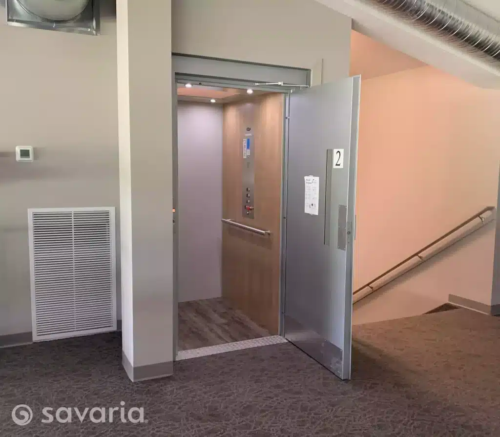 Savaria Prolift Open Vertical Platform Lift with wooden interior and number "2" on the door, in a carpeted hallway.