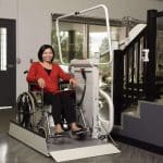 Woman using a Savaria Omega Inclined Platform Lift wheelchair model beside a staircase indoors.