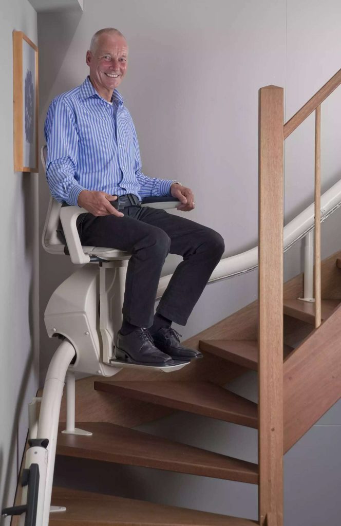 Man sitting on a Savaria Stairfriend 23 stairlift beside a wooden staircase, smiling at the camera.