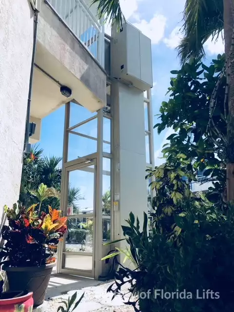 Savaria Enclosed Outdoor Residential 1504 Vertical Platform Lift beside a white building surrounded by plants.