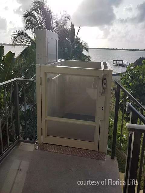 An outdoor view of Savaria V-1504 Vertical Platform Lift on a balcony overlooking water and palm trees.