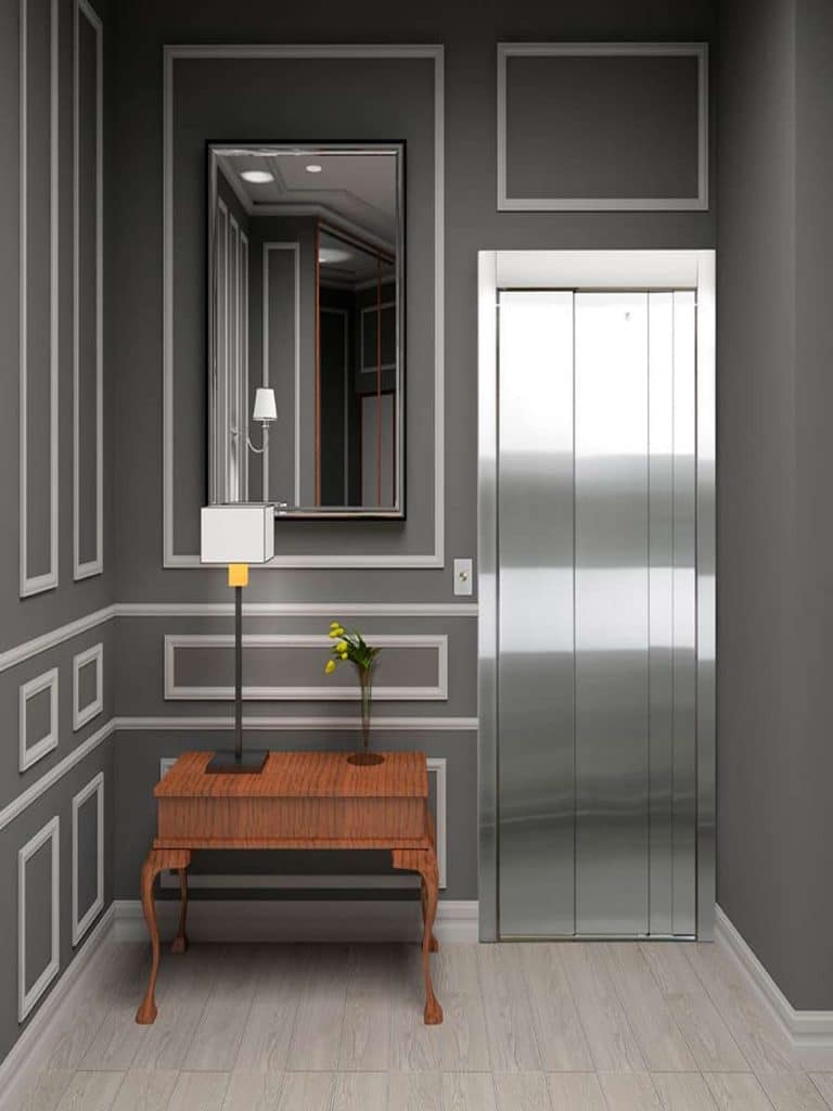 Savaria Zenith Elevator Lobby With Gray Walls, Wooden Table, Table Lamp and Flowers on it and Large Mirror Above the Table.