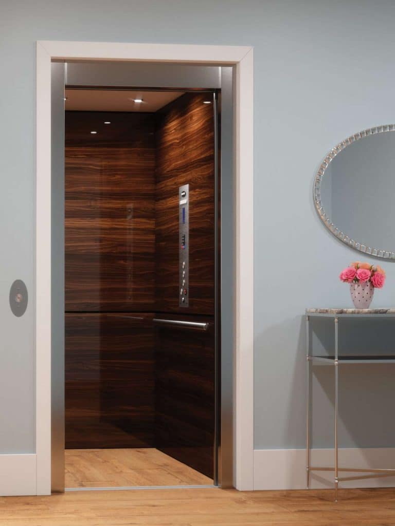 Savaria Zenith Open Elevator with Dark Wood Interior Next to Glass Table Holding Pink Flowers and an Oval Mirror on a Light Blue Wall.