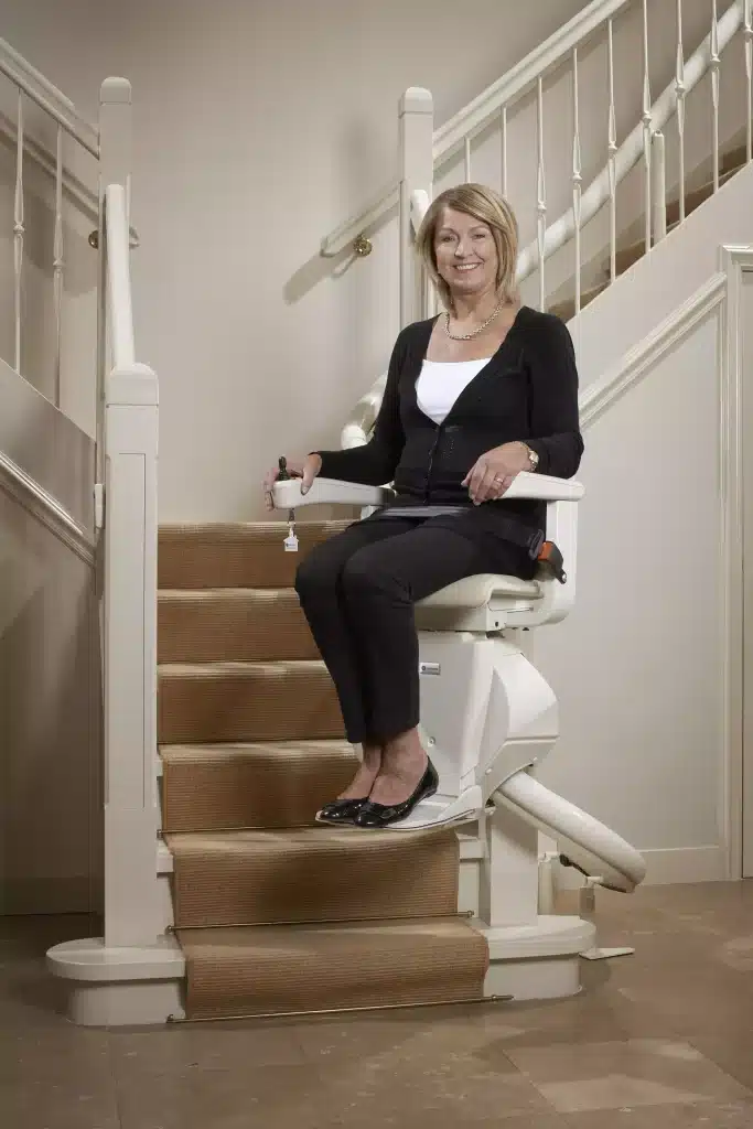 A woman sitting on Handicare Freecurved Stairlift at the bottom of a carpeted staircase holding control buttons.