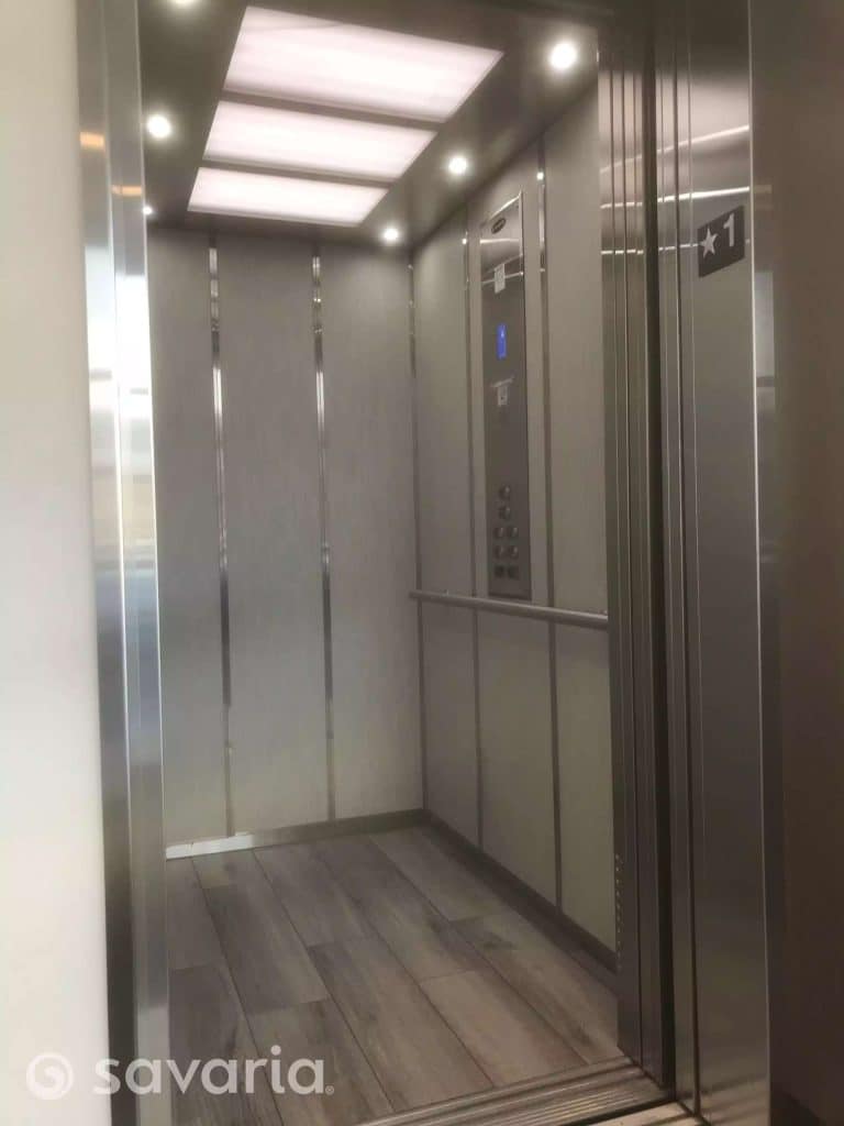 Interior of a modern Savaria Orion17 Commercial elevator with gray walls, metallic accents, wood-patterned floor and bright ceiling lights.