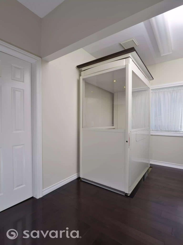 Savaria Telecab Modern White Home Lift Closed Doors With Glass Panels Installed in Corner Of Room With Dark Hardwood Flooring.