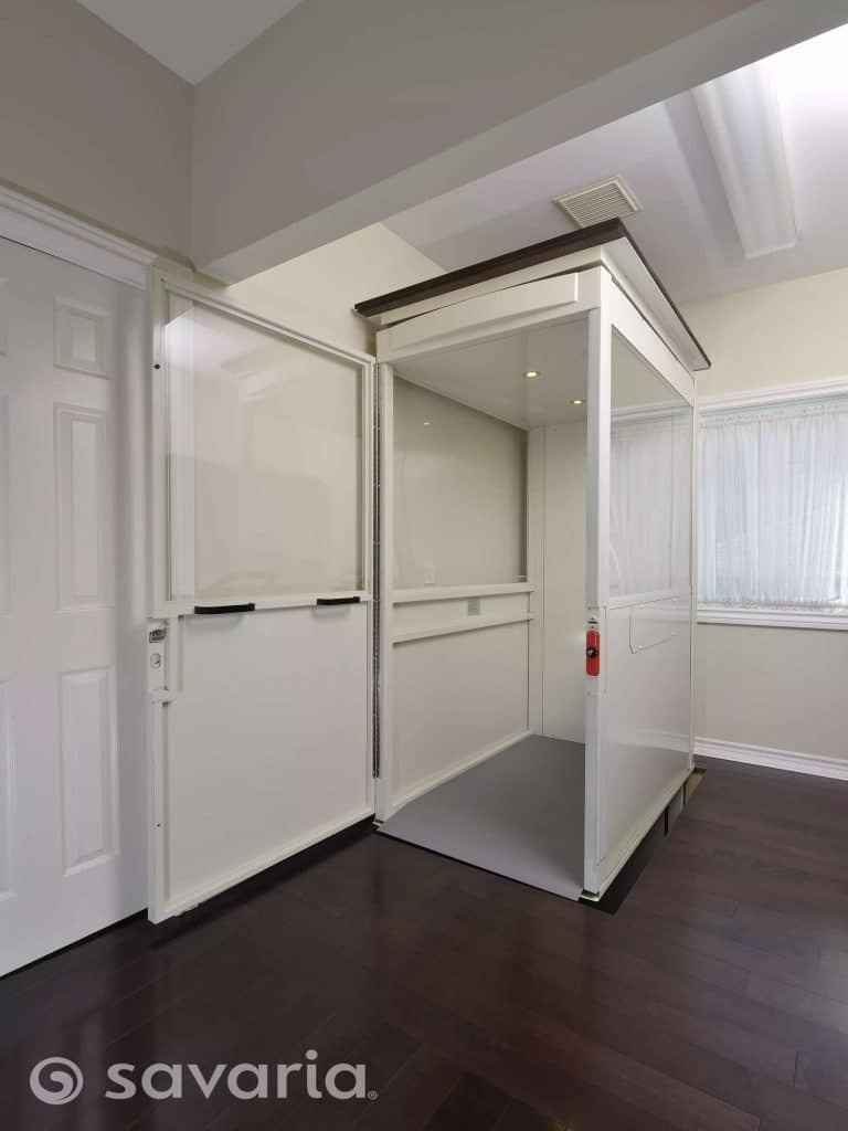 Savaria Telecab Modern White Home Lift Open Doors With Glass Panels Installed in Corner Of Room With Dark Hardwood Flooring.