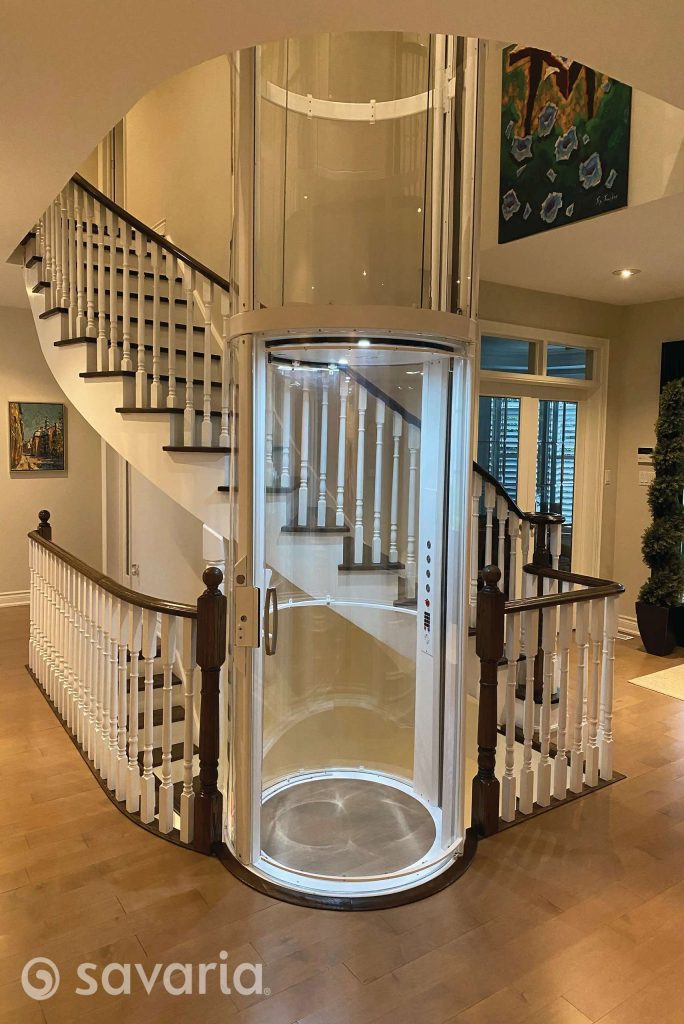 Savaria Vuelift Mini Modern cylindrical glass elevator surrounded by a wooden spiral staircase in a home interior.