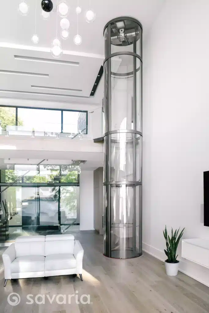 Modern Living Space With Glass Cylindrical Savaria Vuelift Mini Elevator and White Sofa and Large Windows.