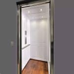 Savaria MDF Material Modern Home Elevator with white paneled walls, recessed lights, wooden floor, and a metallic control panel. Exterior has a gray wall with button panel.