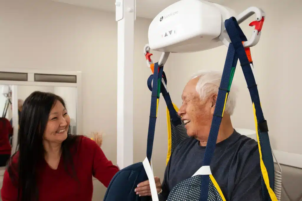 AP Series AP-450 Ceiling Lift, A woman standing next to an older man showing safe and comfortable mobility support in a home care setting.