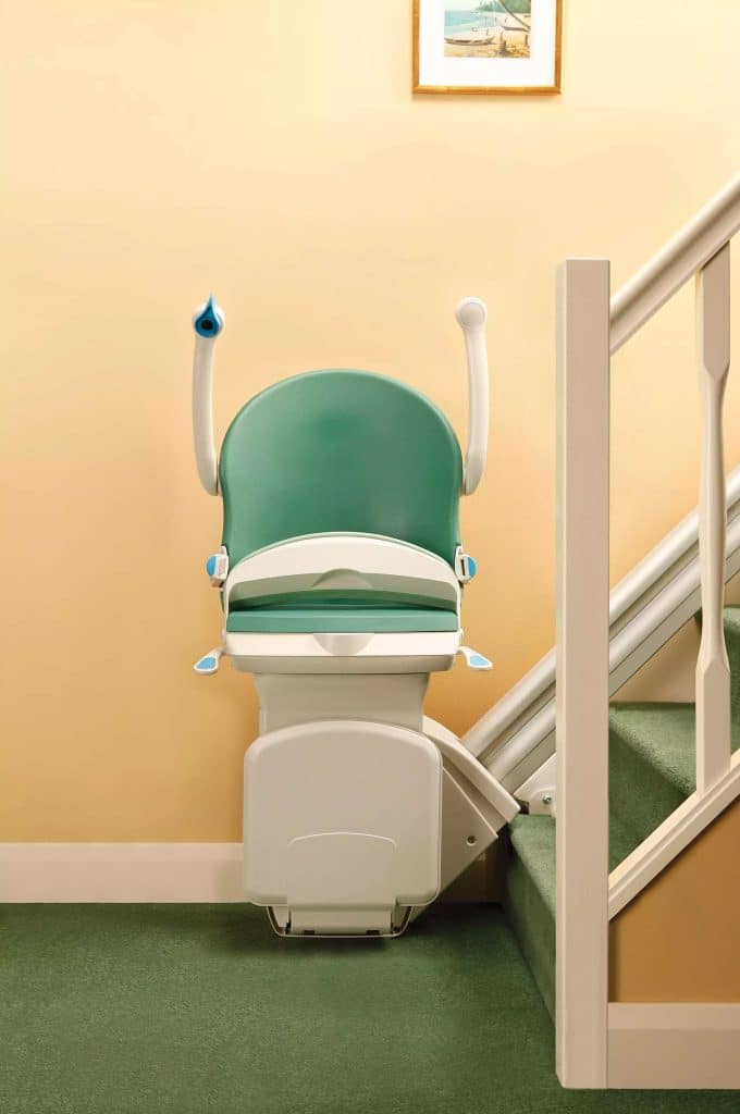 Handicare 1000 Straight Stairlift with a green seat and white armrests installed at the base of a home staircase, with beige walls and a picture above.