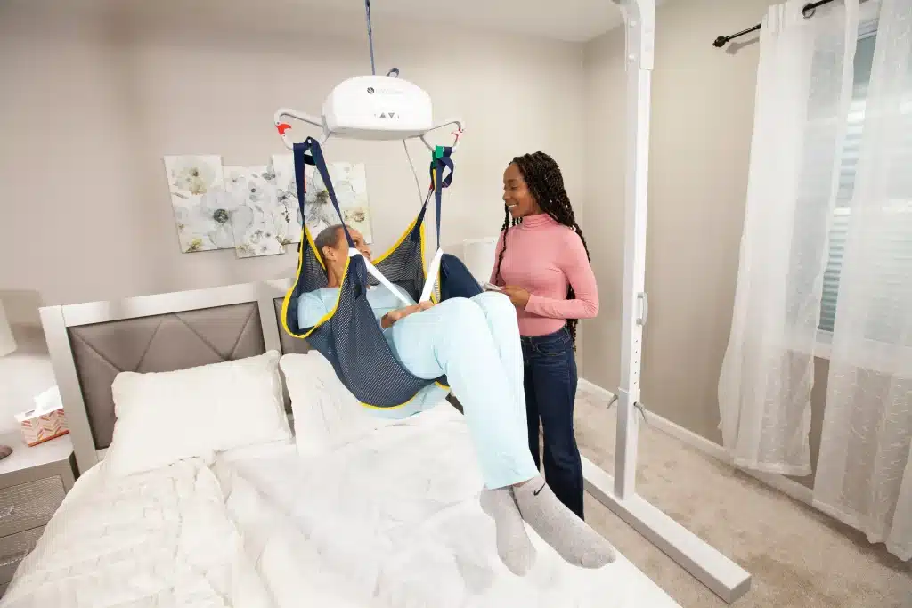 A woman operates Handicare P440 ceiling lift assisting a person in a sling over a bed in a bedroom.