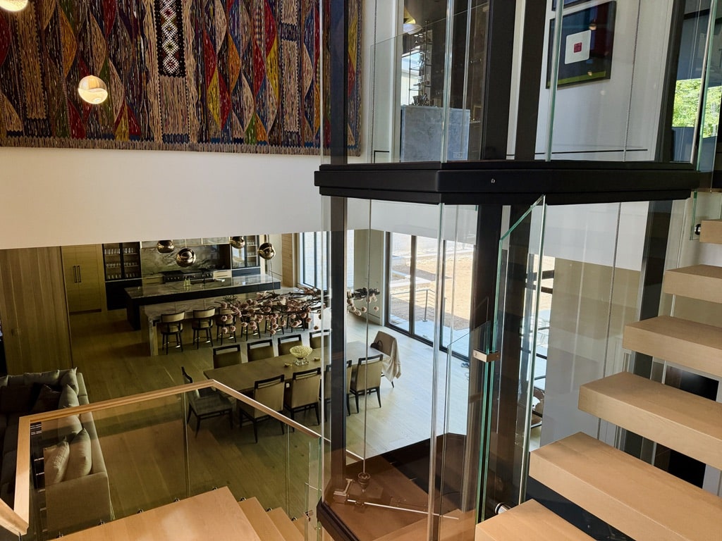 Interior view of a modern Savaria Vuelift glass elevator, wooden staircase and colorful tapestry.