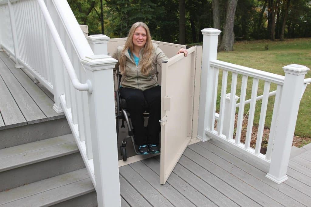 A woman in a Bruno VPL wheelchair inside an outdoor platform lift on a wooden deck with white railings.