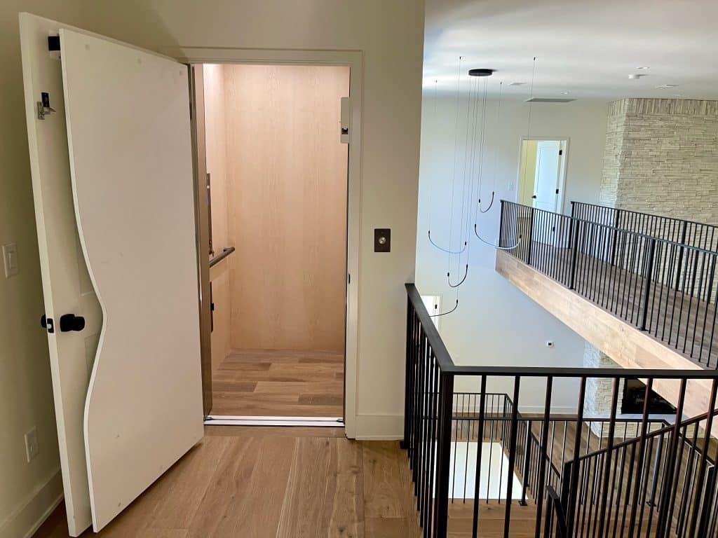 A modern hallway with a wavy-edged Sparta door opening to an elevator, featuring wood flooring and metal railings.